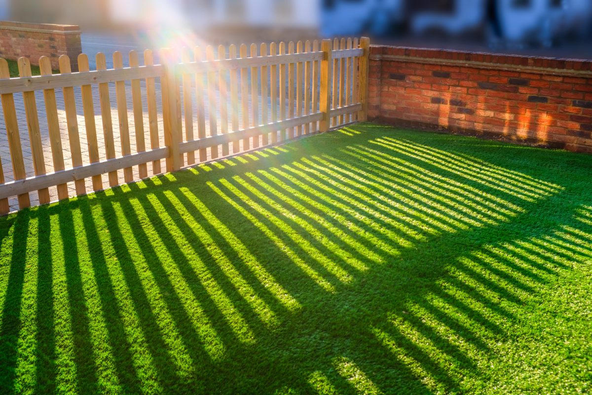 sunshine creating lens flare through a wooden picket fence in a front yard, front garden with artifical grass as a lawn and a red brick perimiter wall.

