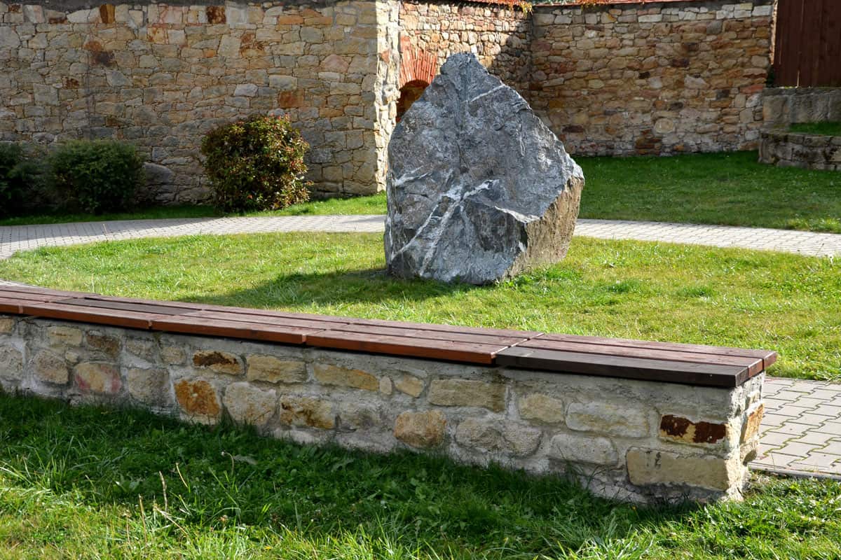 sun lit photo of a park with a large rock on the center, huge stone, wall bench on the side
