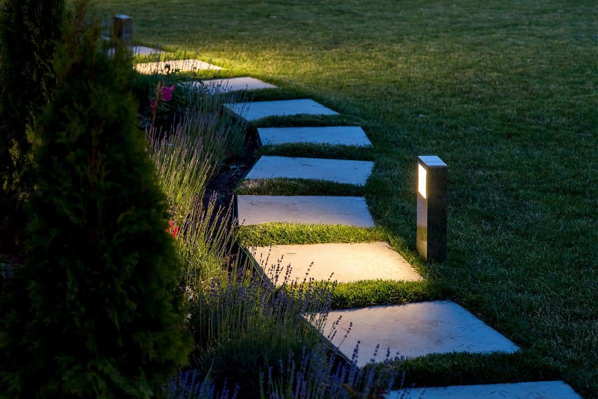 Marble path of square tiles illuminated by a lantern glowing with a warm light in a backyard garden with a flower bed and a lawn copy space. — Photo
