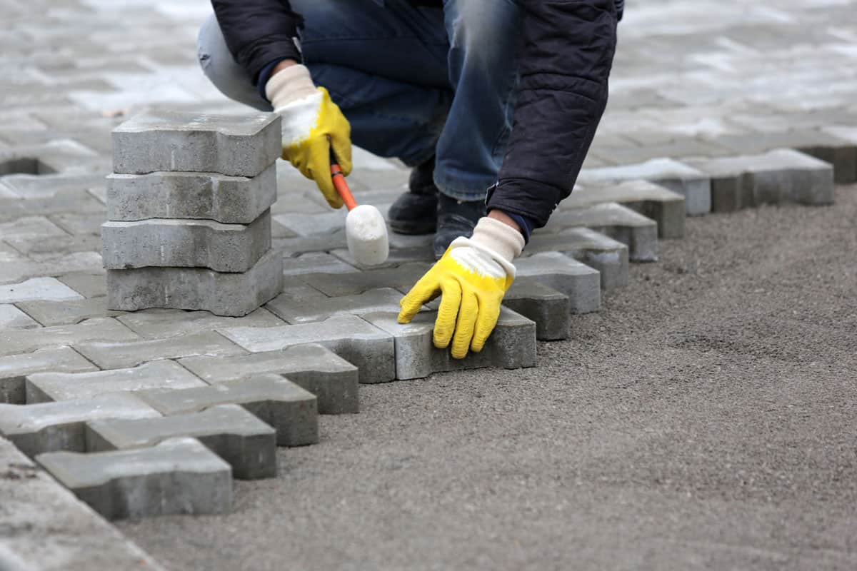 man wearing yellow safety gloves, holding rubber mallet, installing concrete pavers